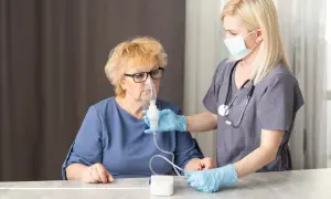Pielęgniarka pomaga starszej kobiecie z maską inhalacyjną. Dowiedz się, jak zrobić inhalacje z soli fizjologicznej bez inhalatora.