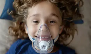 Uśmiechnięty chłopiec z kręconymi włosami ma na twarzy maskę inhalatora. Warto wiedzieć, co wlać do inhalatora, by pomóc dziecku.