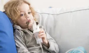 Chłopiec z blond włosami korzysta z inhalatora. Po inhalacji może być potrzebne oklepywanie dziecka, by ułatwić odkrztuszanie wydzieliny.