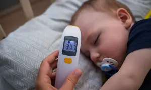 Dziecko śpi z termometrem wskazującym 37.7°C. Rodzic przygotowuje zimne okłady na gorączkę u dziecka.