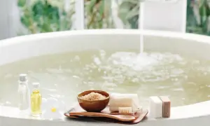 Relaks w wannie z wodą solankową, obok kosmetyki: olejki, peeling, mydła i kwiat.