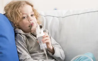 Chłopiec z blond włosami korzysta z inhalatora. Po inhalacji może być potrzebne oklepywanie dziecka, by ułatwić odkrztuszanie wydzieliny.