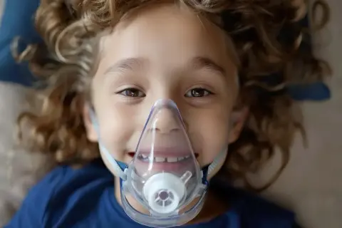 Uśmiechnięty chłopiec z kręconymi włosami ma na twarzy maskę inhalatora. Warto wiedzieć, co wlać do inhalatora, by pomóc dziecku.