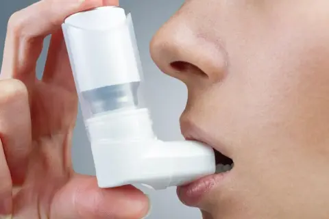 Dłoń trzyma biały inhalator na astmę, który osoba wkłada do ust.