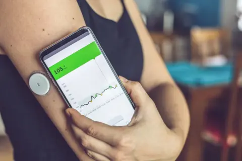 Osoba sprawdza na telefonie wykres poziomu glukozy. Glukometr pokazuje 105 mg/dL, ale nie widać, czy wynik jest prawidłowy.