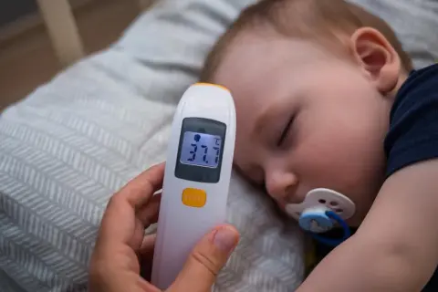 Dziecko śpi z termometrem wskazującym 37.7°C. Rodzic przygotowuje zimne okłady na gorączkę u dziecka.