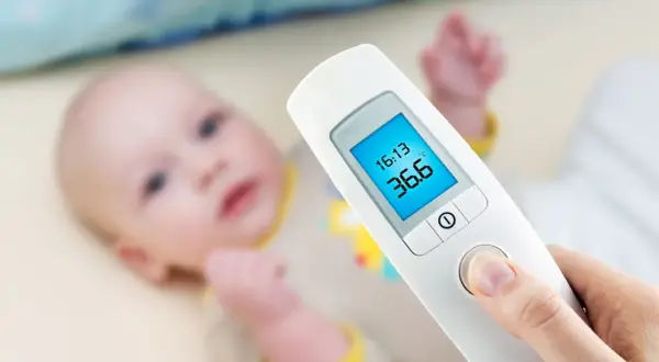 Termometr dla niemowlaka pokazuje 36.6°C. Dziecko leży w tle.