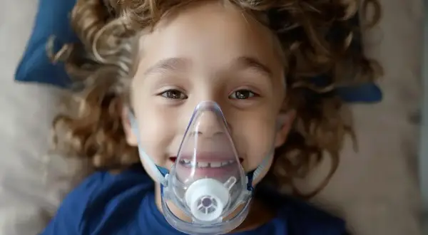 Uśmiechnięty chłopiec z kręconymi włosami ma na twarzy maskę inhalatora. Warto wiedzieć, co wlać do inhalatora, by pomóc dziecku.