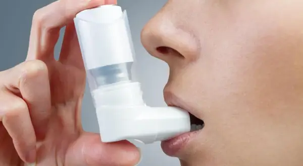 Dłoń trzyma biały inhalator na astmę, który osoba wkłada do ust.
