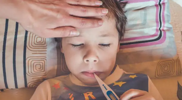 Dziecko z gorączką i wymiotami leży w łóżku, z termometrem w ustach. Ręka rodzica na czole sprawdza temperaturę.