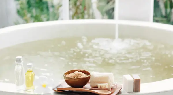 Relaks w wannie z wodą solankową, obok kosmetyki: olejki, peeling, mydła i kwiat.