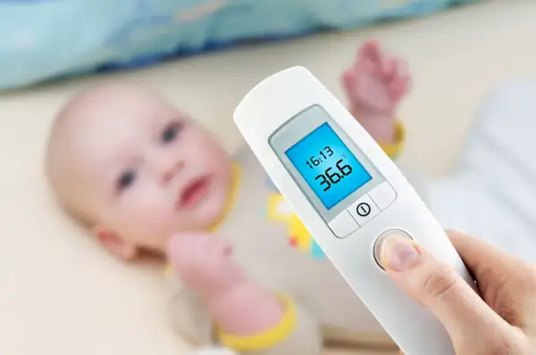 Termometr dla niemowlaka pokazuje 36.6°C. Dziecko leży w tle.